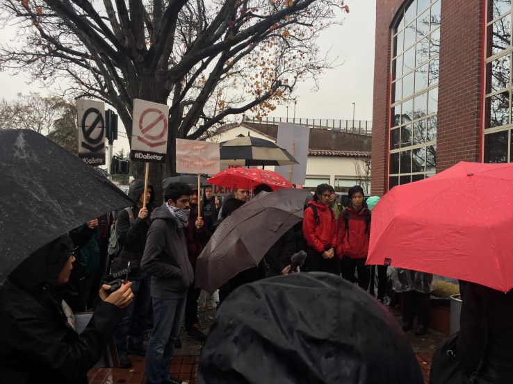 Dozens of protestors huddle in the rain outside Palantir.